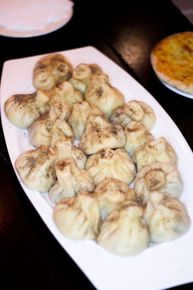 A white platter of steamed dumplings, sprinkled with black pepper, resting on a dark wooden table.