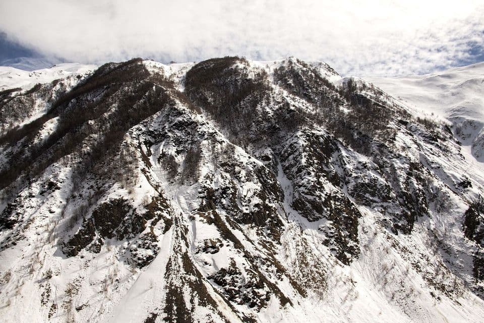 A steep, rugged mountain covered in snow, with patches of rock and bare trees visible under a partly cloudy sky.