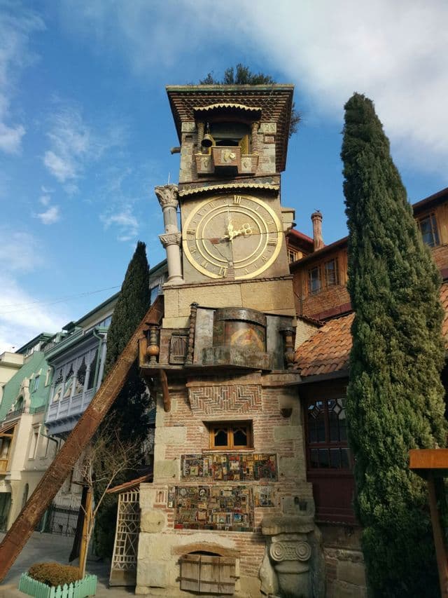 Una torre dell'orologio stravagante e pendente con una grande facciata dorata si erge su una strada cittadina accanto a un alto cipresso.