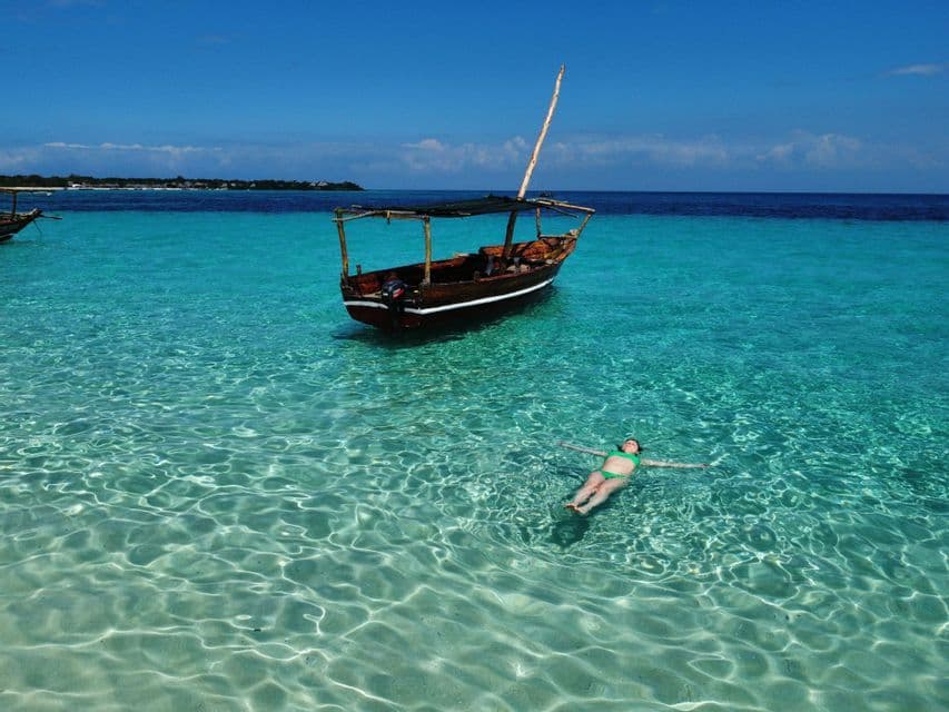 A person floats on their back in clear turquoise water next to a traditional wooden boat under a blue sky.