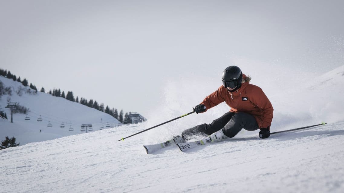 Uno sciatore con giacca arancione e casco nero scende una pista innevata, sollevando spruzzi di neve, con una seggiovia sullo sfondo.