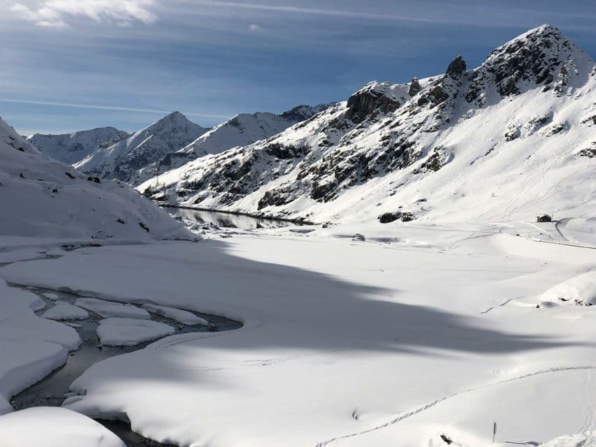 Un lago parzialmente ghiacciato giace in una valle circondata da montagne innevate sotto un cielo azzurro e limpido.