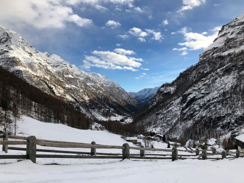 Una staccionata di legno delimita un campo innevato che si affaccia su un piccolo villaggio in una valle montana sotto un cielo parzialmente nuvoloso