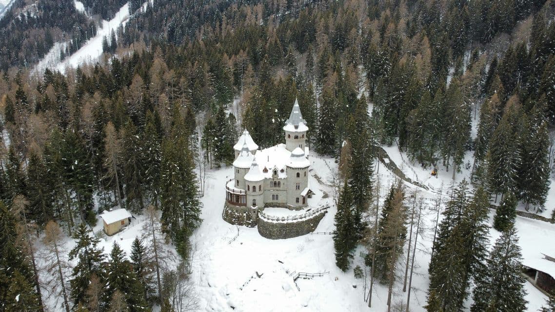 Una veduta aerea di un castello di pietra con torrette e tetti innevati, circondato da una fitta foresta invernale.