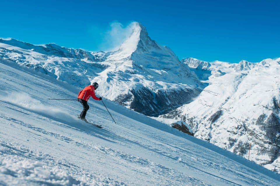 Uno sciatore con giacca rossa scende un pendio montano innevato, con una vetta prominente visibile contro un cielo azzurro chiaro.