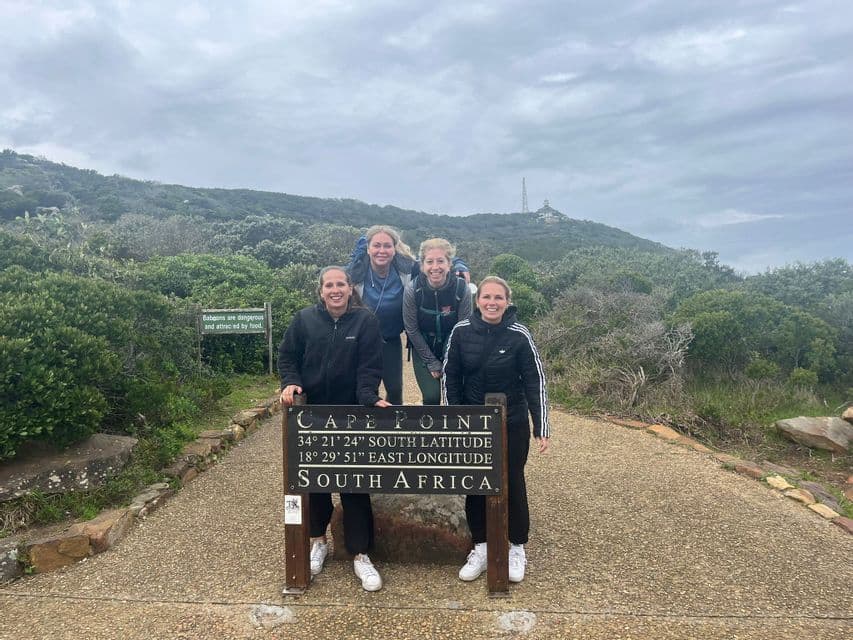Eine WeRoad-Gruppenreise mit vier Frauen, die am Schild von Cape Point, Südafrika, auf einem Pfad durch eine hügelige Landschaft posieren.