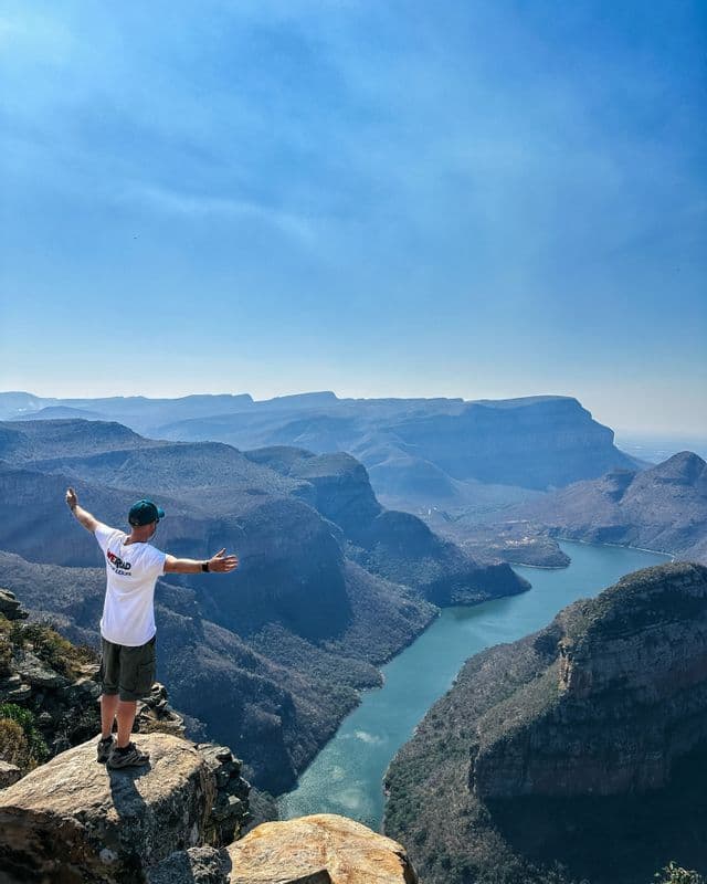 Ein Mann auf einem WeRoad-Trip steht mit ausgestreckten Armen auf einer felsigen Klippe und überblickt eine weite Schlucht und einen sich windenden Fluss.