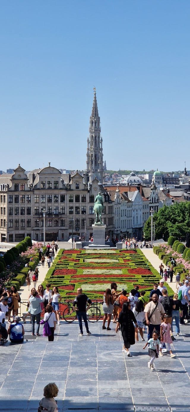 Passeggiano in un giardino formale in una piazza cittadina, con edifici storici e un'alta guglia sullo sfondo.