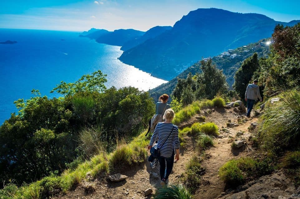 A WeRoad group trip hiking on a sunlit coastal trail overlooking a blue sea and mountains.