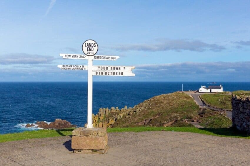 Il cartello bianco di Land's End con le distanze per varie località, affacciato su una scogliera verde e il mare blu.