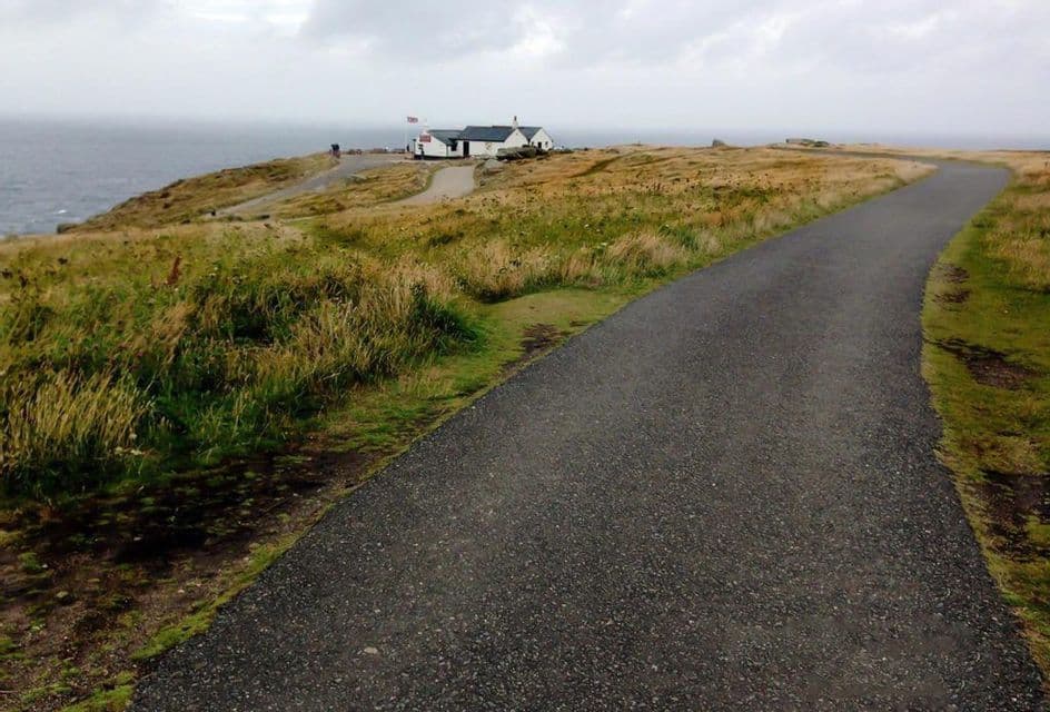 Un sentiero lastricato si snoda lungo una scogliera erbosa verso un edificio bianco a picco sul mare sotto un cielo coperto.