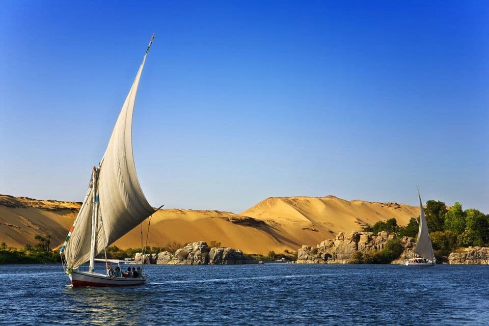 Une felouque traditionnelle avec une grande voile blanche glisse sur un fleuve bleu, devant des dunes de sable du désert et une rive rocheuse.