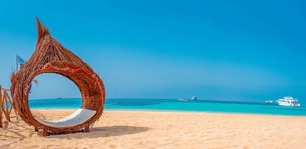 Un transat en osier façon nid, avec un coussin blanc, est installé sur une plage de sable fin, face à des eaux turquoise sous un ciel bleu clair.