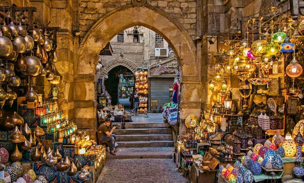 Une vue à travers une arche de pierre, vers une ruelle de marché bordée d'étals proposant de nombreuses lampes et lanternes colorées et illuminées.