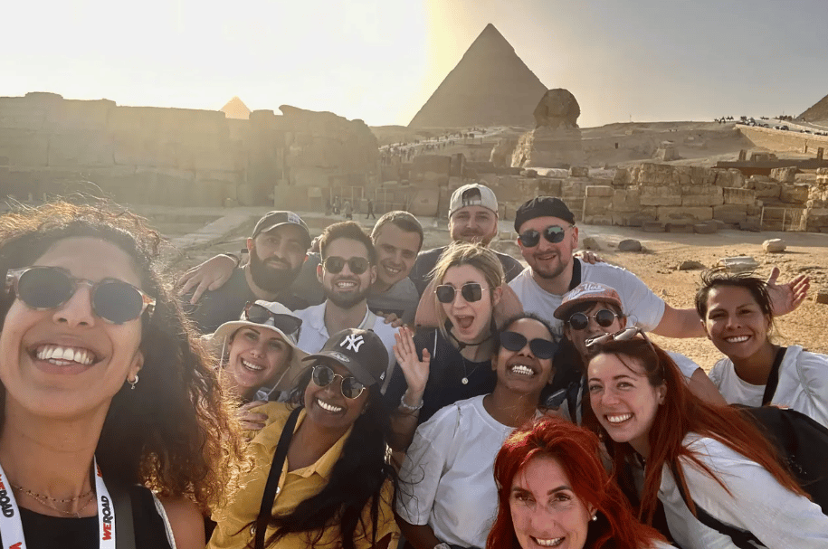 Un groupe WeRoad prend un selfie souriant devant les pyramides et le Sphinx lors d'une journée ensoleillée.