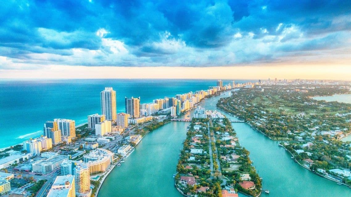 Una vista aerea di una città costiera con alti edifici sul lungomare e isole residenziali verdi separate da un canale turchese.