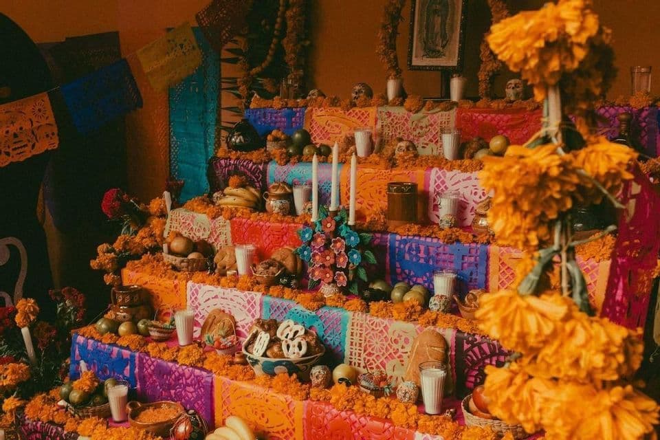 A colorful, multi-tiered Day of the Dead altar decorated with orange marigolds, candles, fruit, bread, and skulls.