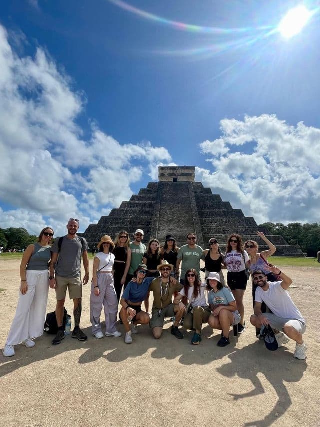 A WeRoad group trip smiles for a photo in front of a large stone pyramid under a bright, cloudy sky.