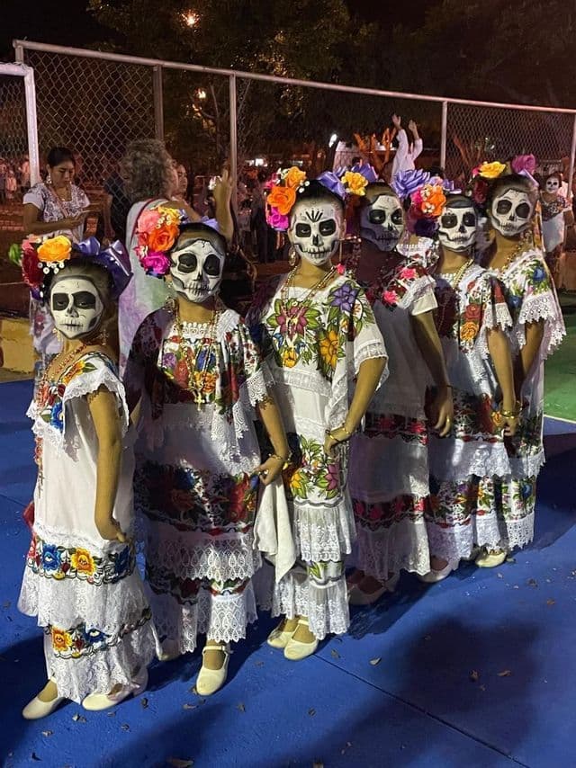 A group of young women with skull face paint and floral headdresses stand in a line wearing traditional embroidered dresses at night.