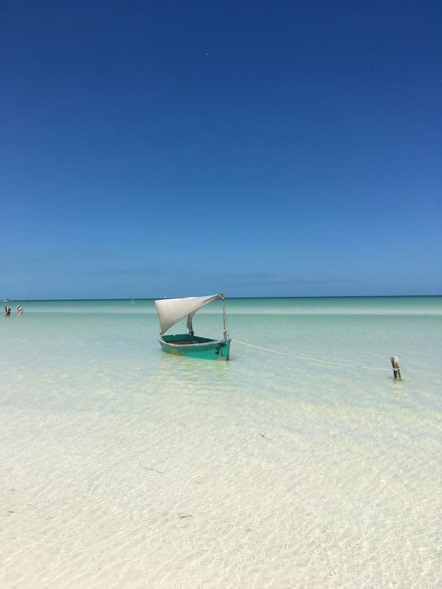 A small green boat with a white canopy is moored in shallow, clear turquoise water under a cloudless blue sky.