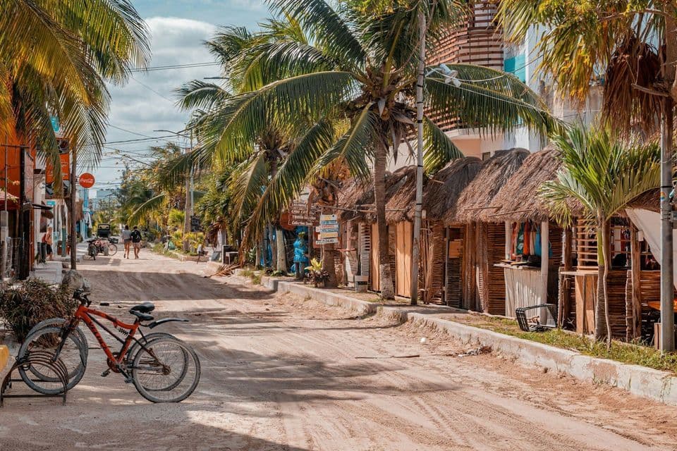 A sandy street in a tropical town lined with palm trees and small shops with thatched roofs, with a red bicycle parked in the foreground.