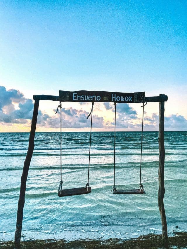 Two empty wooden swings hang from a frame on a beach, facing the ocean and a colorful sky at sunrise.