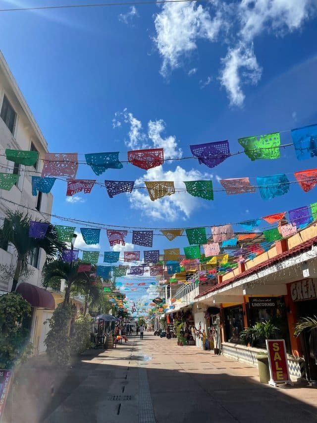 Colorful papel picado banners are strung across a sunlit pedestrian street lined with shops under a blue sky.