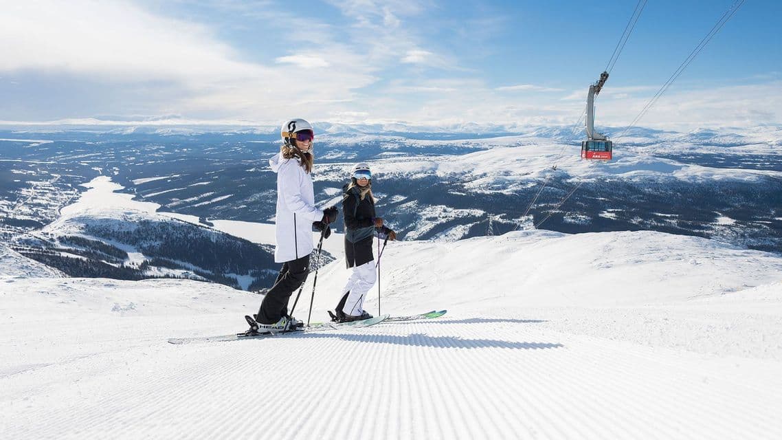 Due donne in tenuta da sci su una pista battuta, con vista su una vasta valle innevata e una funivia rossa sullo sfondo.