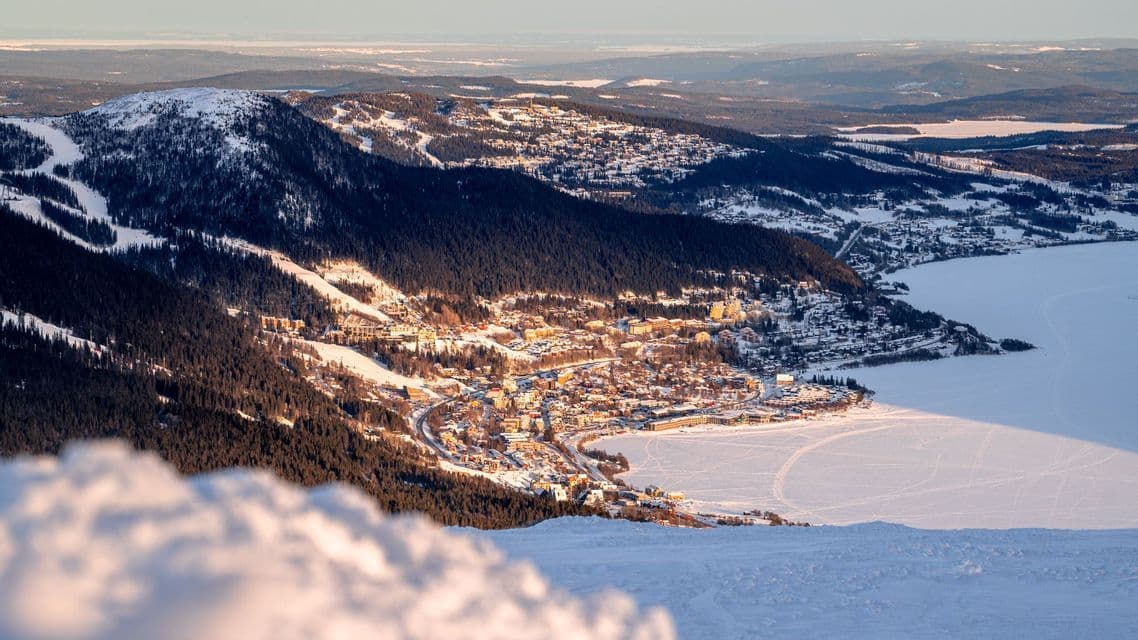 Una vista dall'alto di una città innevata e illuminata dal sole, sulla riva di un lago ghiacciato, circondata da montagne boscose.