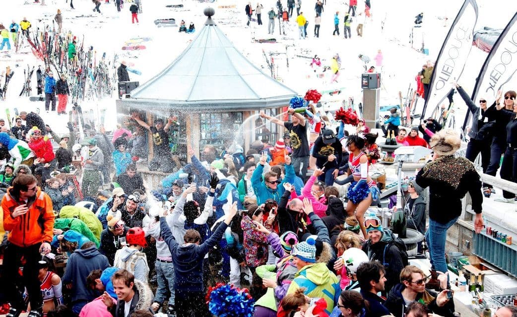 Un viaggio di gruppo WeRoad festeggia a una affollata festa après-ski su una pista innevata, con persone che ballano e spruzzano champagne.