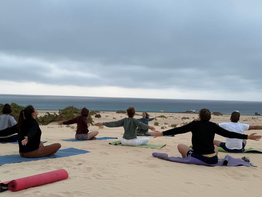 Un voyage de groupe WeRoad pratique le yoga sur des tapis disposés sur une dune de sable, surplombant l'océan sous un ciel nuageux.