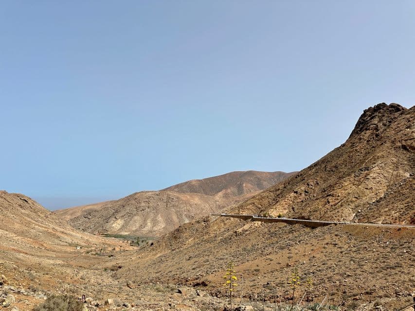 Une route sinueuse traverse une vallée sèche et rocheuse entre des montagnes arides sous un ciel bleu clair.