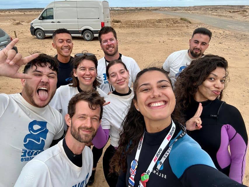 Un groupe WeRoad en voyage prend un selfie dans un espace extérieur sablonneux, avec des participants portant des combinaisons de plongée.