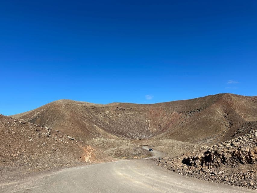 Un chemin de terre serpente à travers un paysage de cratère volcanique rocailleux, avec une voiture au loin sous un ciel bleu clair.