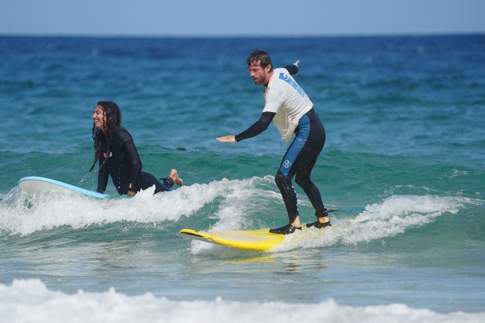 Un homme d'un voyage de groupe WeRoad se tient sur une planche de surf jaune tandis qu'une femme souriante est allongée sur une autre planche dans la mer.