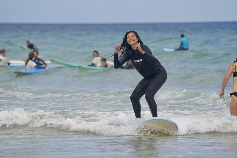 Une femme d'un voyage de groupe WeRoad sourit en surfant sur une vague, avec d'autres personnes en arrière-plan.