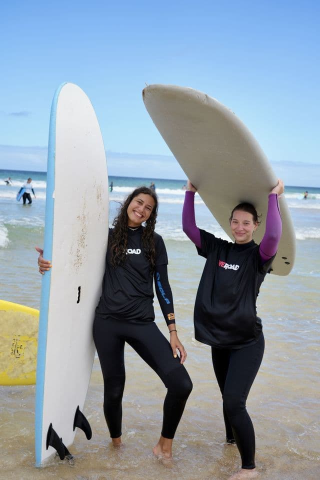 Deux femmes d'un voyage de groupe WeRoad sourient et posent avec des planches de surf, debout dans l'eau peu profonde sur la plage.