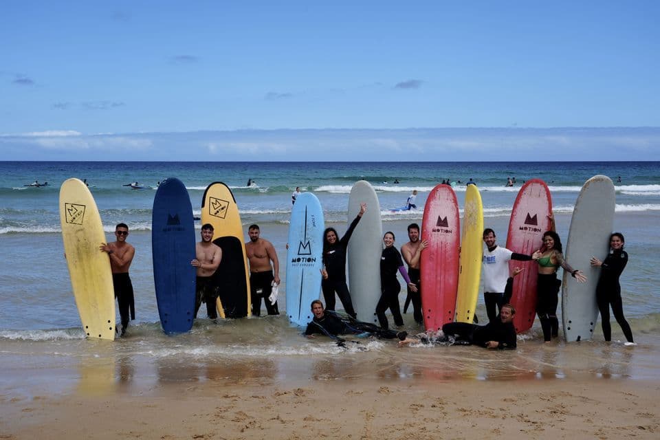 Un groupe WeRoad posant avec des planches de surf colorées dans l'eau peu profonde d'une plage sous un ciel bleu.