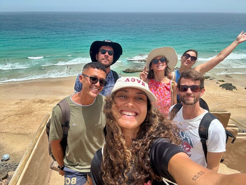 Un voyage de groupe WeRoad de six personnes souriant pour un selfie avec vue sur une plage de sable et l'océan turquoise.