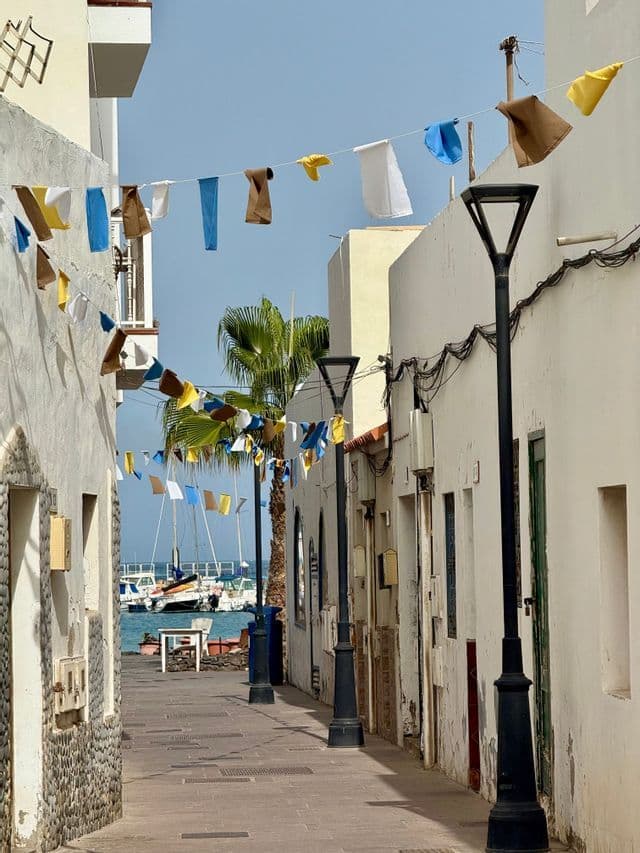 Une ruelle étroite entre des bâtiments blancs, décorée de guirlandes colorées, mène à un port avec des bateaux sur l'eau.