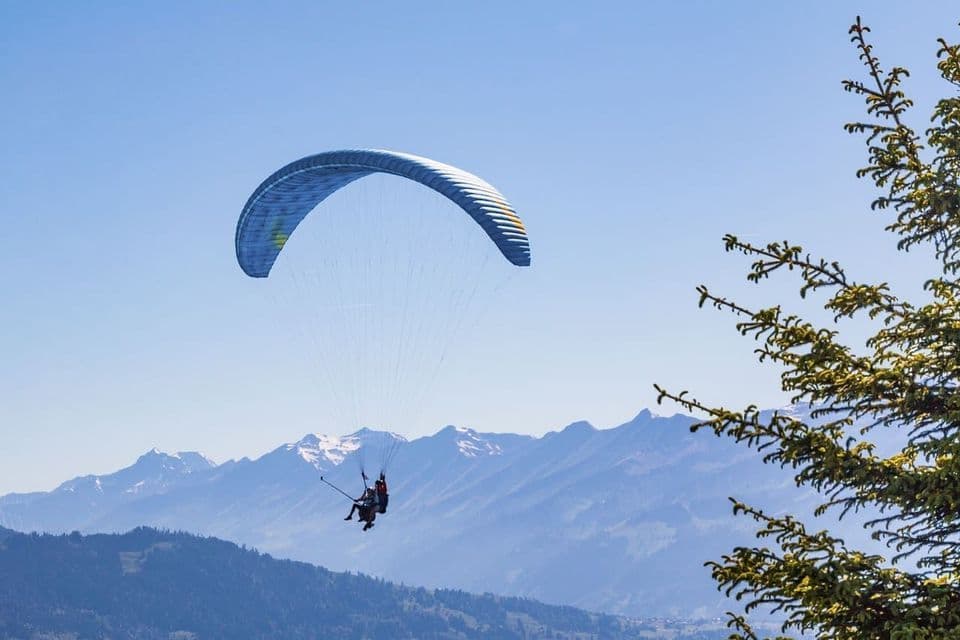 Deux personnes en parapente biplace s'élèvent au-dessus d'une vaste chaîne de montagnes aux sommets enneigés sous un ciel bleu clair.