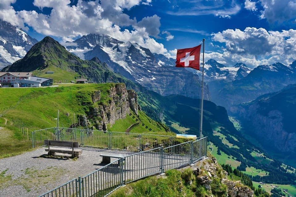 Un drapeau suisse flotte à un point de vue avec un banc, surplombant des pentes verdoyantes et des montagnes enneigées sous un ciel nuageux.