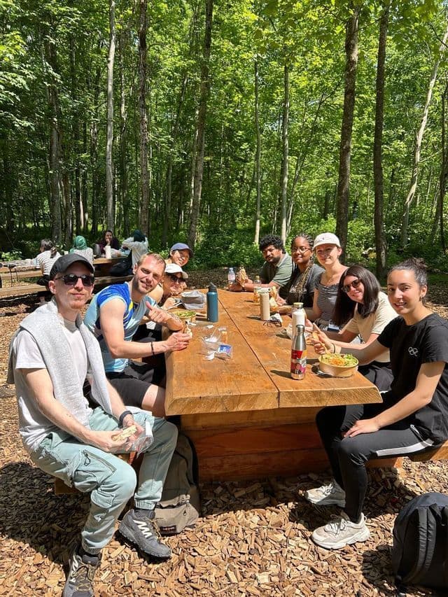 Un groupe WeRoad déjeune ensemble à une longue table de pique-nique en bois dans une forêt ensoleillée.