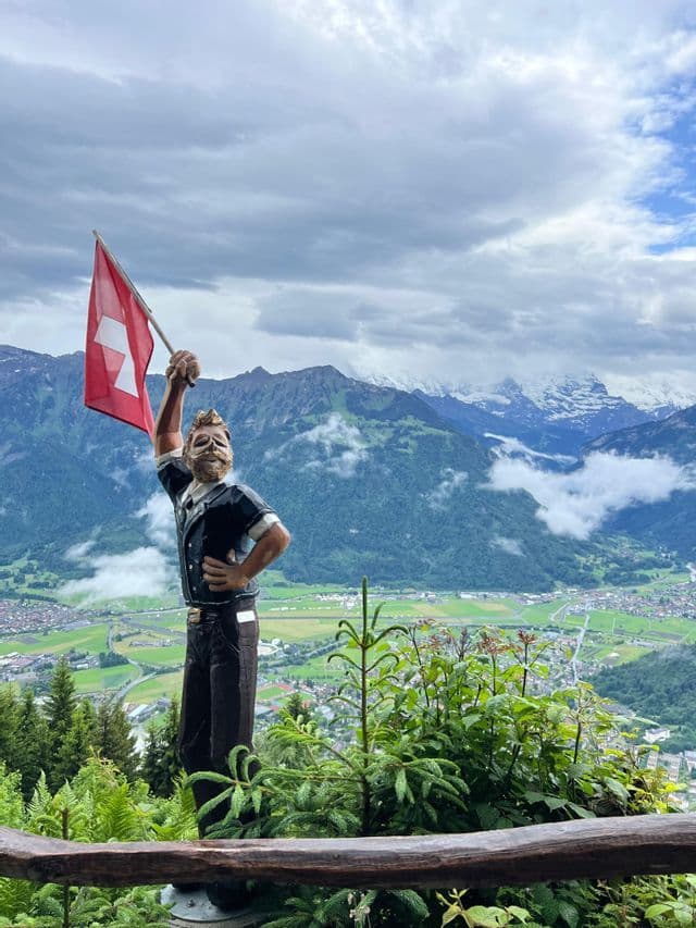 Une statue en bois d'un homme tient un drapeau suisse, surplombant une vallée verdoyante avec des montagnes enneigées en arrière-plan.