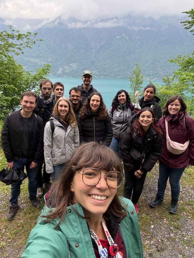 Une femme en veste verte prend un selfie souriant avec un groupe de voyage WeRoad, avec un lac turquoise et des montagnes boisées en arrière-plan.
