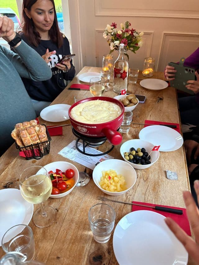 Un groupe WeRoad en voyage, réuni autour d'une table en bois, dégustant une fondue au fromage suisse avec du pain, des olives et du vin.