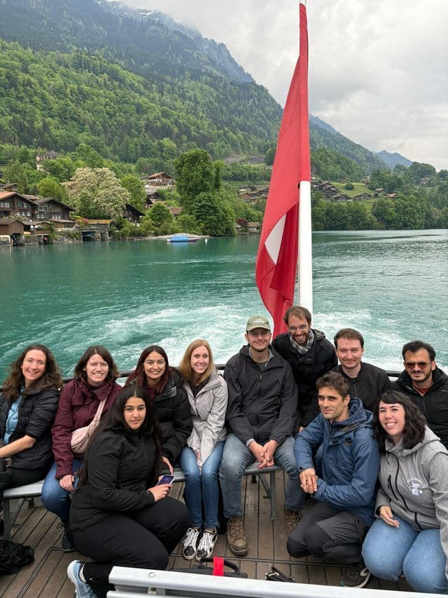 Un groupe WeRoad pose pour une photo sur un bateau naviguant sur un lac turquoise avec des montagnes boisées et un village en arrière-plan.