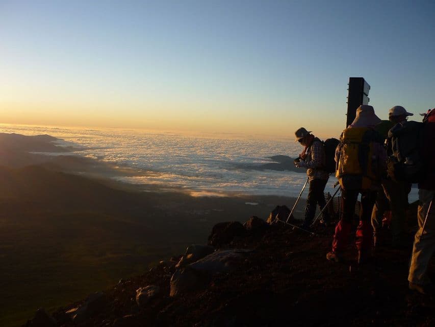 Un gruppo WeRoad di escursionisti con zaini si erge su una cima rocciosa all'alba, ammirando un vasto mare di nuvole.