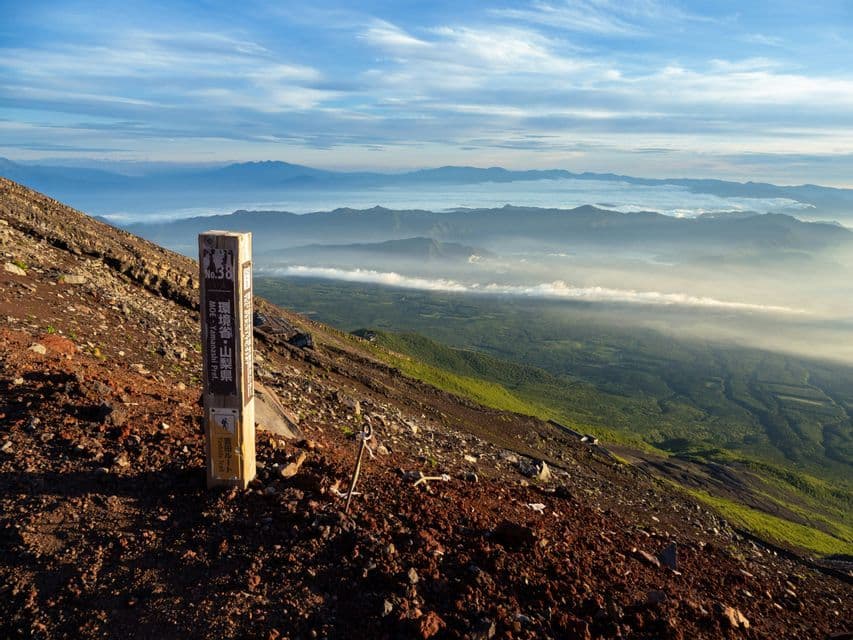 Un segnavia in legno su un fianco roccioso di montagna, affacciato su una vasta valle piena di nuvole e lontane catene montuose.