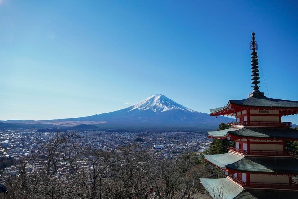 Una pagoda giapponese rossa si affaccia su una città con il Monte Fuji innevato che si erge sullo sfondo contro un cielo azzurro chiaro.